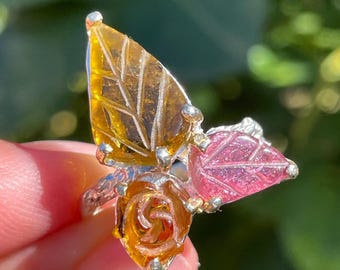 May include: A silver ring featuring a floral design with a rose and two leaves. The leaves are a translucent yellow and pink, and the rose is a translucent orange. The ring is set against a blurred green background.