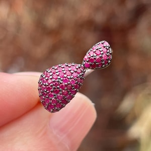 Puede incluir: Primer plano de un pequeño colgante de metal oscuro con un grupo de pequeñas gemas redondas de color rosa brillante. El colgante tiene forma de lágrima con una parte superior más pequeña y redondeada. Las gemas están muy juntas.