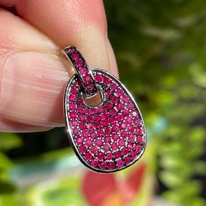 May include: Oval-shaped pendant with a silver-colored frame, filled with numerous small, vibrant red gemstones. The pendant has a loop at the top for a chain. The background is blurred, with green foliage and a hint of red flowers.