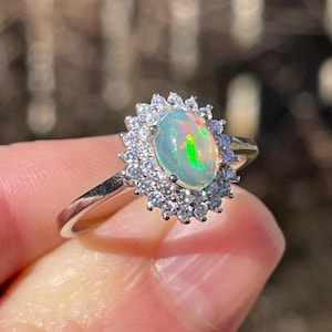 May include: A silver ring with an oval opal gemstone, surrounded by small, clear gemstones. The opal has flashes of green and orange. The ring is held up against a blurred outdoor background. The ring is a piece of jewellery.