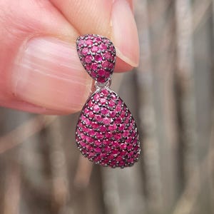 May include: A silver earring with a teardrop design, encrusted with small, bright pink gemstones. The earring is held between fingers, highlighting its detailed design and colour.
