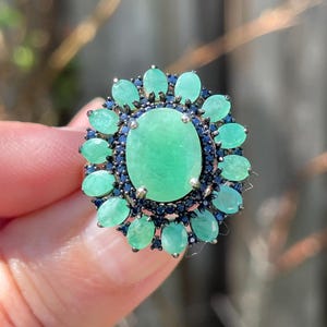 Puede incluir: Un anillo plateado con una gran piedra preciosa verde claro de talla ovalada en el centro, rodeada de piedras verdes claras ovaladas más pequeñas y piedras azul oscuro. El anillo se sostiene sobre un fondo borroso.