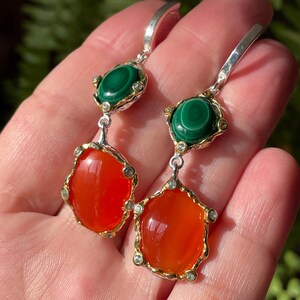 May include: A pair of dangle earrings featuring a silver hook, a green malachite stone, and a large orange carnelian stone. The stones are set in gold-colored metal with small, clear accent stones. The earrings are displayed against a blurred green background.