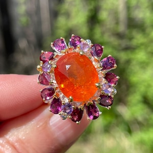 Puede incluir: Un anillo vibrante con una gran piedra preciosa naranja ovalada en el centro, rodeada por un halo de piedras preciosas más pequeñas y multicolores en tonos púrpura y rosa. El anillo tiene un engaste dorado.