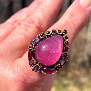 May include: A large, ornate ring featuring a large, teardrop-shaped pink gemstone. The gemstone is surrounded by smaller, dark red and purple gemstones set in a gold-toned metal frame. The ring is a statement piece of jewelry.