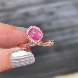 May include: A rose gold ring with a pink oval gemstone set in a delicate, ornate setting. The ring features a twisted band with a textured finish.
