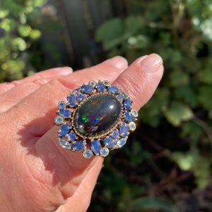 May include: A large, ornate ring featuring a dark, oval-shaped opal with iridescent flashes. The opal is surrounded by a halo of blue and light blue gemstones set in a gold-toned metal band. The ring is displayed on a hand.