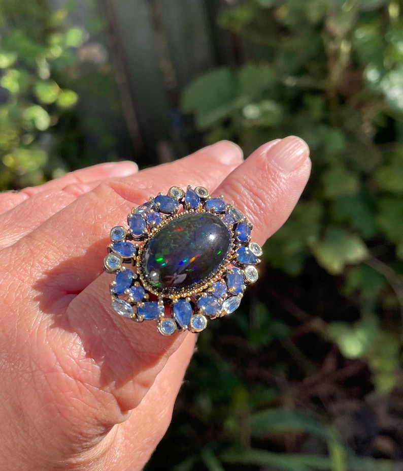 May include: A large, ornate ring featuring a dark, oval-shaped opal with iridescent flashes. The opal is surrounded by a halo of blue and light blue gemstones set in a gold-toned metal band. The ring is displayed on a hand.