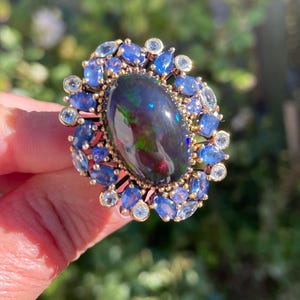 May include: A gold ring featuring a large, oval black opal gemstone with iridescent flashes of color. The opal is surrounded by a halo of blue sapphires and smaller, round, clear gemstones. The ring is set against a blurred green background.