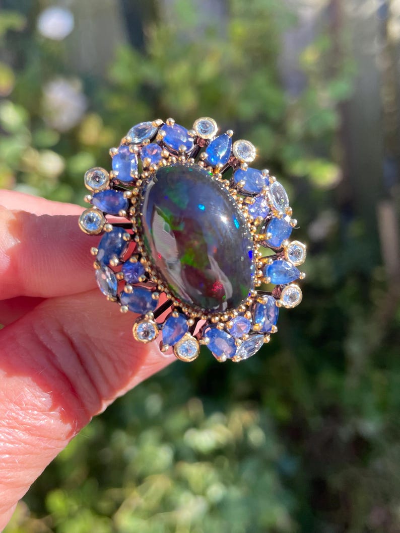 May include: A gold ring featuring a large, oval black opal gemstone with iridescent flashes of color. The opal is surrounded by a halo of blue sapphires and smaller, round, clear gemstones. The ring is set against a blurred green background.