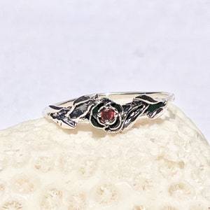Może przedstawiać: Srebrny pierścionek z kwiatowym wzorem i czerwonym kamieniem.