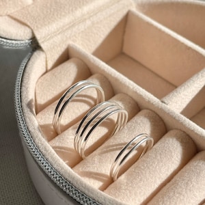 May include: Three silver rings of different sizes are displayed in a white ring box with beige velvet lining.