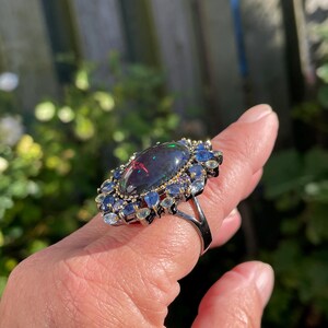 May include: A silver ring featuring a large, oval-shaped black opal gemstone with iridescent flashes of color. The opal is surrounded by a halo of smaller blue and clear gemstones, creating a striking contrast. The ring is set against a natural outdoor background.