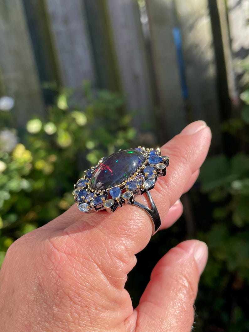 May include: A silver ring featuring a large, oval-shaped black opal gemstone with iridescent flashes of color. The opal is surrounded by a halo of smaller blue and clear gemstones, creating a striking contrast. The ring is set against a natural outdoor background.