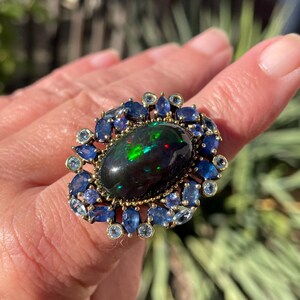 May include: A gold ring featuring a large, oval black opal with vibrant green and red flashes. The opal is surrounded by a halo of blue sapphires and smaller, light blue gemstones.