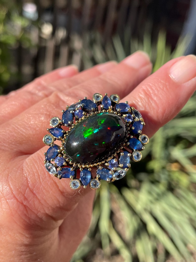 May include: A gold ring featuring a large, oval black opal with vibrant green and red flashes. The opal is surrounded by a halo of blue sapphires and smaller, light blue gemstones.