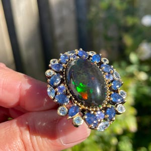 May include: A gold-toned ring featuring a large, oval black opal with vibrant color flashes. The opal is surrounded by a halo of blue and clear gemstones, creating a striking contrast. The ring is held up against a blurred green background.