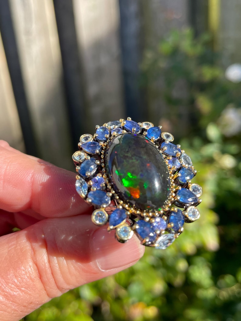 May include: A gold-toned ring featuring a large, oval black opal with vibrant color flashes. The opal is surrounded by a halo of blue and clear gemstones, creating a striking contrast. The ring is held up against a blurred green background.