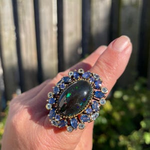 May include: A large, ornate ring featuring a dark oval-shaped gemstone at the center, surrounded by smaller blue and clear gemstones. The ring has a gold-colored band and setting, creating a striking contrast with the dark central stone and the blue accents.