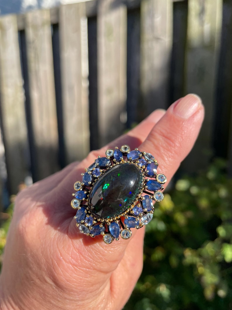 May include: A large, ornate ring featuring a dark oval-shaped gemstone at the center, surrounded by smaller blue and clear gemstones. The ring has a gold-colored band and setting, creating a striking contrast with the dark central stone and the blue accents.