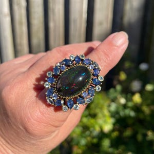 May include: A large, ornate ring featuring a dark oval gemstone at the center, surrounded by smaller blue and light blue stones. The ring has a gold-colored setting and is displayed on a finger against a blurred outdoor background.