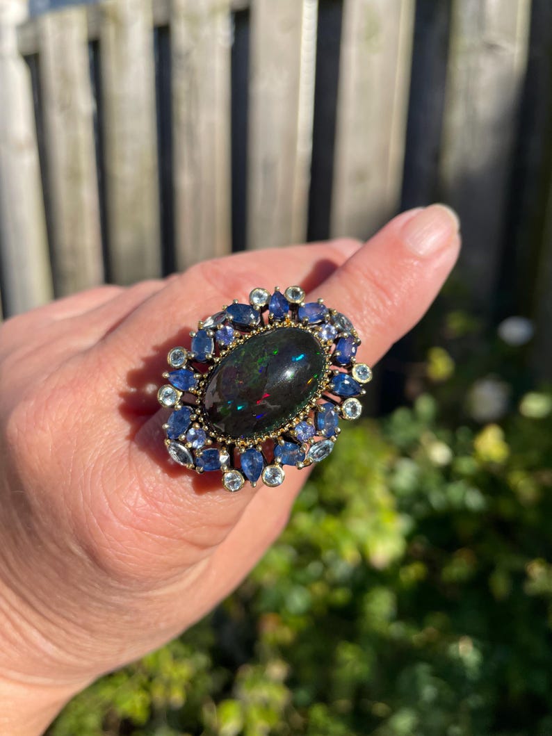 May include: A large, ornate ring featuring a dark oval gemstone at the center, surrounded by smaller blue and light blue stones. The ring has a gold-colored setting and is displayed on a finger against a blurred outdoor background.