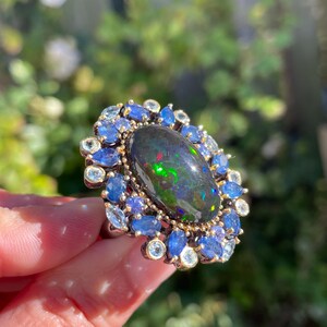 May include: An oval-shaped ring featuring a large, dark opal with vibrant green and red flashes. The opal is surrounded by a halo of blue sapphires and smaller, clear gemstones, set in a gold-toned metal.