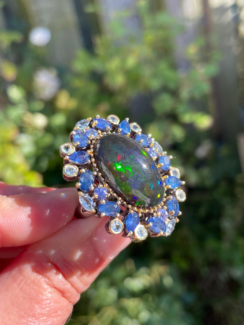 May include: An oval-shaped ring featuring a large, dark opal with vibrant green and red flashes. The opal is surrounded by a halo of blue sapphires and smaller, clear gemstones, set in a gold-toned metal.