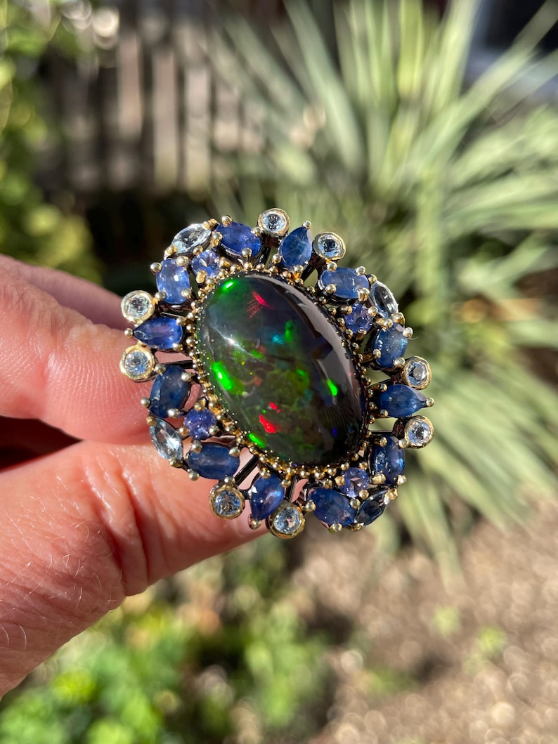 Exquisite large genuine Ethiopian pinfire black opal artisan ring. 

US size: 8
Main gemstone: Ethiopian black opal
Other gemstones: blue sapphire and sky blue topaz
Main stone: 21 x 14mm (14ct)
Treatment: heated
Material: 925 sterling silver
Plating: black rhodium and 18K yellow gold
Face ring: 35 x 28mm
Weight: 14.5g (gem(s)+setting)