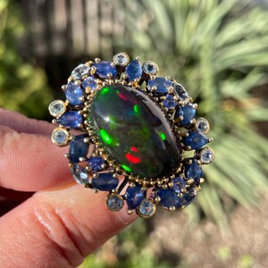 May include: An oval-shaped ring featuring a large, iridescent black opal gemstone. The opal is surrounded by a halo of blue sapphires and smaller, light blue gemstones, set in a gold-toned metal. The ring's design is intricate and eye-catching.