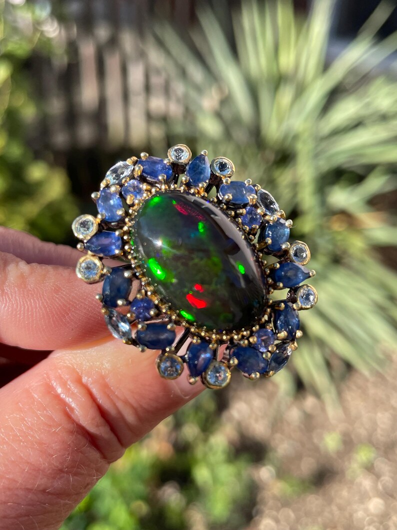 May include: An oval-shaped ring featuring a large, iridescent black opal gemstone. The opal is surrounded by a halo of blue sapphires and smaller, light blue gemstones, set in a gold-toned metal. The ring's design is intricate and eye-catching.