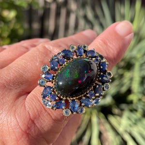 May include: An ornate ring featuring a large, oval black opal gemstone with iridescent flashes. The opal is surrounded by a halo of blue sapphires and smaller, light blue gemstones, set in a gold-toned metal band. The ring is a statement piece.