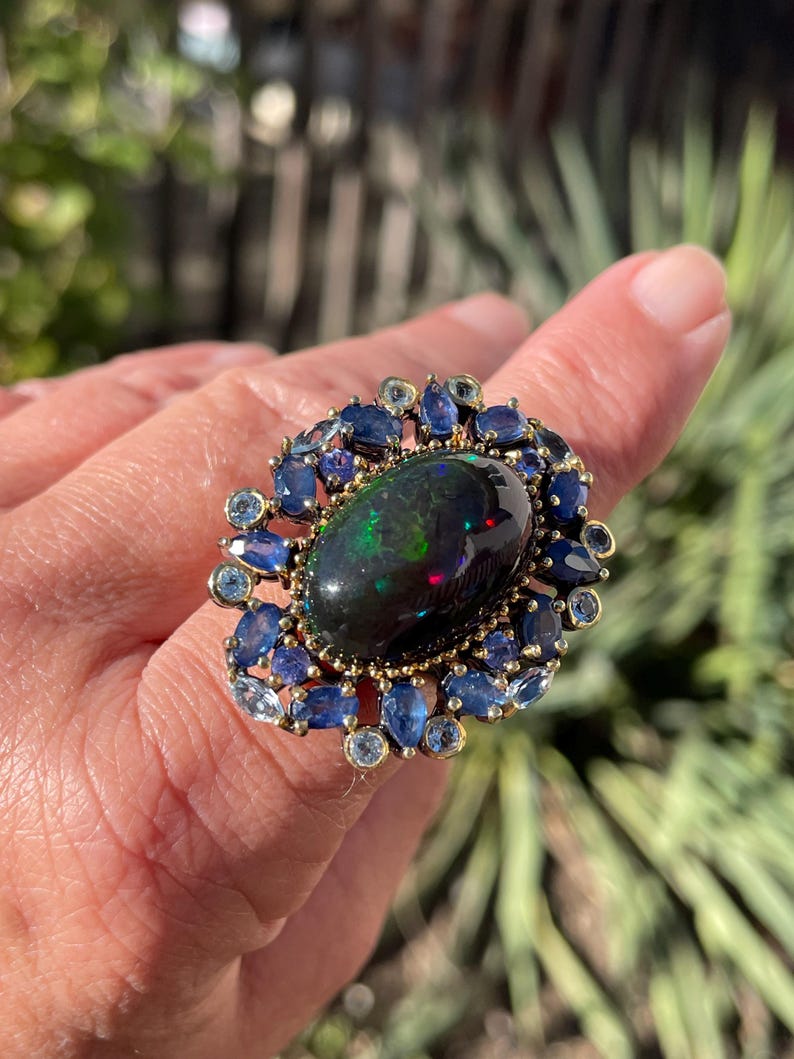 May include: An ornate ring featuring a large, oval black opal gemstone with iridescent flashes. The opal is surrounded by a halo of blue sapphires and smaller, light blue gemstones, set in a gold-toned metal band. The ring is a statement piece.