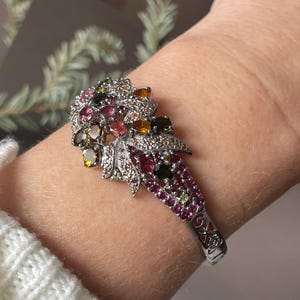 Puede incluir: Una pulsera de plata con un diseño floral que presenta una variedad de piedras preciosas de colores, incluyendo rosa, verde y amarillo.