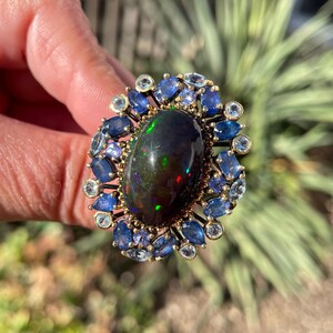 May include: A gold ring featuring a large, oval black opal gemstone with iridescent flashes. The opal is surrounded by a halo of blue sapphires and smaller, round gemstones. The ring is held up against a blurred green background.