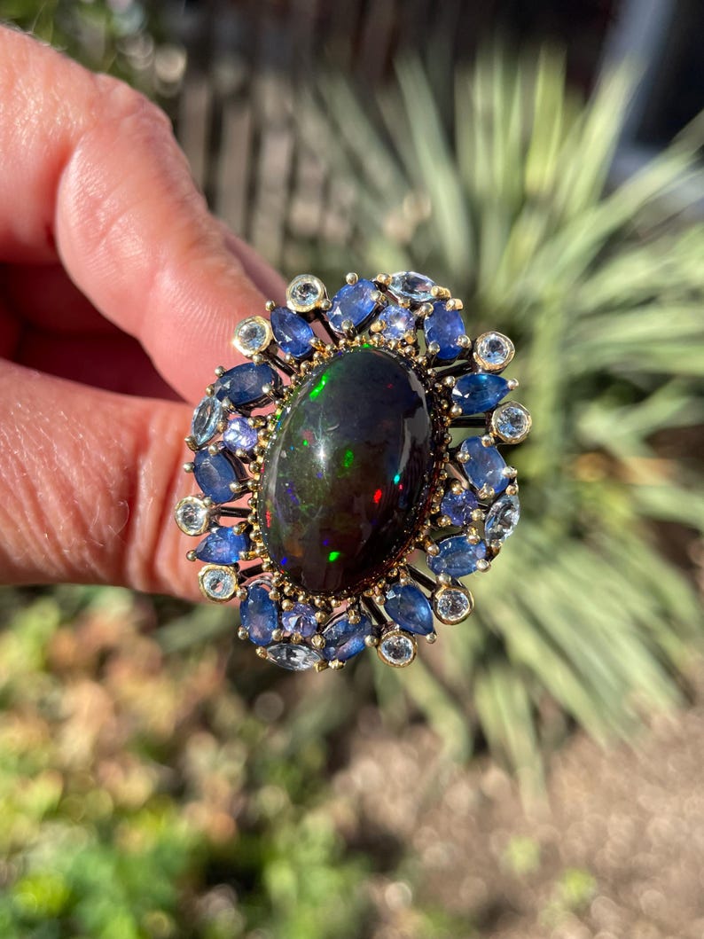 May include: A gold ring featuring a large, oval black opal gemstone with iridescent flashes. The opal is surrounded by a halo of blue sapphires and smaller, round gemstones. The ring is held up against a blurred green background.