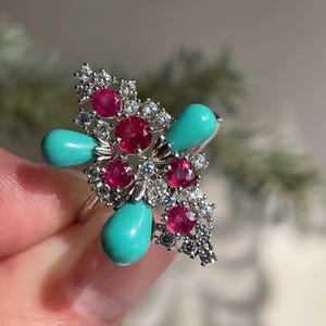 May include: A silver ring featuring a floral design with vibrant gemstones. The ring showcases bright red rubies, sparkling white diamonds, and teardrop-shaped turquoise stones. The ring is a statement piece.