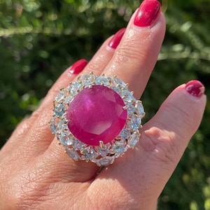 May include: A large, round, faceted magenta-pink gemstone set in a gold-toned metal ring. The central stone is surrounded by a halo of smaller, light blue, oval-cut stones arranged in a floral pattern, creating a wide, decorative statement piece.
