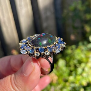 May include: A large, ornate ring featuring a dark, oval-shaped gemstone with iridescent flashes of color. The central stone is surrounded by a halo of smaller blue and clear gemstones, set in a dark metal band. The ring is held up against a blurred green background.