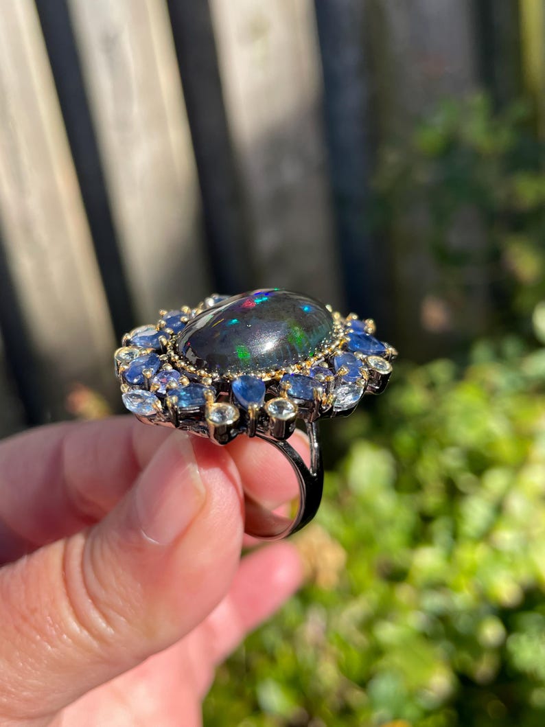 May include: A large, ornate ring featuring a dark, oval-shaped gemstone with iridescent flashes of color. The central stone is surrounded by a halo of smaller blue and clear gemstones, set in a dark metal band. The ring is held up against a blurred green background.