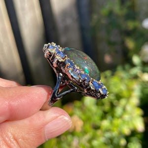 May include: A silver ring featuring a large, oval black opal gemstone with flashes of color. The opal is surrounded by a halo of small, blue gemstones. The ring is set against a blurred green background.