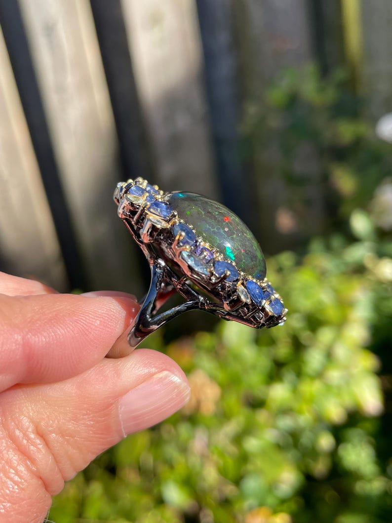 May include: A silver ring featuring a large, oval black opal gemstone with flashes of color. The opal is surrounded by a halo of small, blue gemstones. The ring is set against a blurred green background.