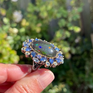 May include: A silver ring featuring a large, oval black opal with iridescent flashes of color. The opal is surrounded by a halo of blue and light blue gemstones, creating a striking contrast against the dark opal.