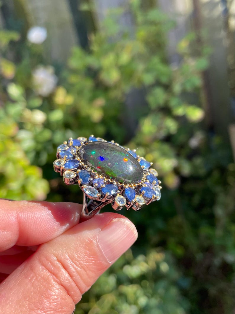 May include: A silver ring featuring a large, oval black opal with iridescent flashes of color. The opal is surrounded by a halo of blue and light blue gemstones, creating a striking contrast against the dark opal.