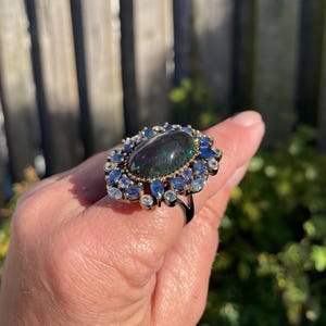 May include: A large, ornate ring featuring a dark, oval-shaped gemstone at its center, surrounded by smaller blue and clear gemstones. The ring has a black band and gold accents, creating a striking contrast. The ring is a statement piece.