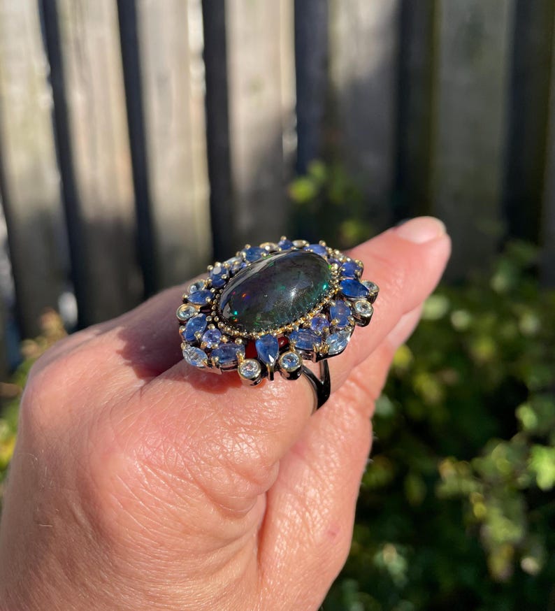 May include: A large, ornate ring featuring a dark, oval-shaped gemstone at its center, surrounded by smaller blue and clear gemstones. The ring has a black band and gold accents, creating a striking contrast. The ring is a statement piece.