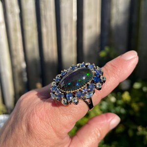 May include: A large, oval-shaped black opal ring with a halo of blue and clear gemstones. The ring has a dark metal band and is set against a natural outdoor background. The opal displays a play of color, with flashes of green and blue.
