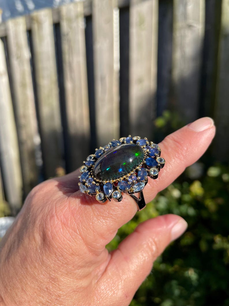 May include: A large, oval-shaped black opal ring with a halo of blue and clear gemstones. The ring has a dark metal band and is set against a natural outdoor background. The opal displays a play of color, with flashes of green and blue.