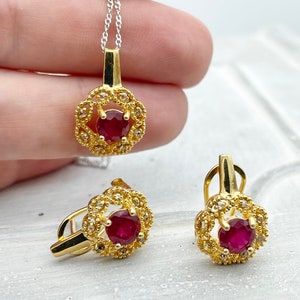 Puede incluir: Un conjunto de collar y pendientes de oro con una piedra preciosa de rubí rojo rodeada de piedras blancas más pequeñas en un diseño floral.