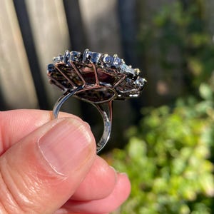May include: A silver ring with a large, ornate setting. The setting is a cluster of small, dark stones surrounding larger, clear stones. The ring is held up by a person's fingers, with a blurred green background.